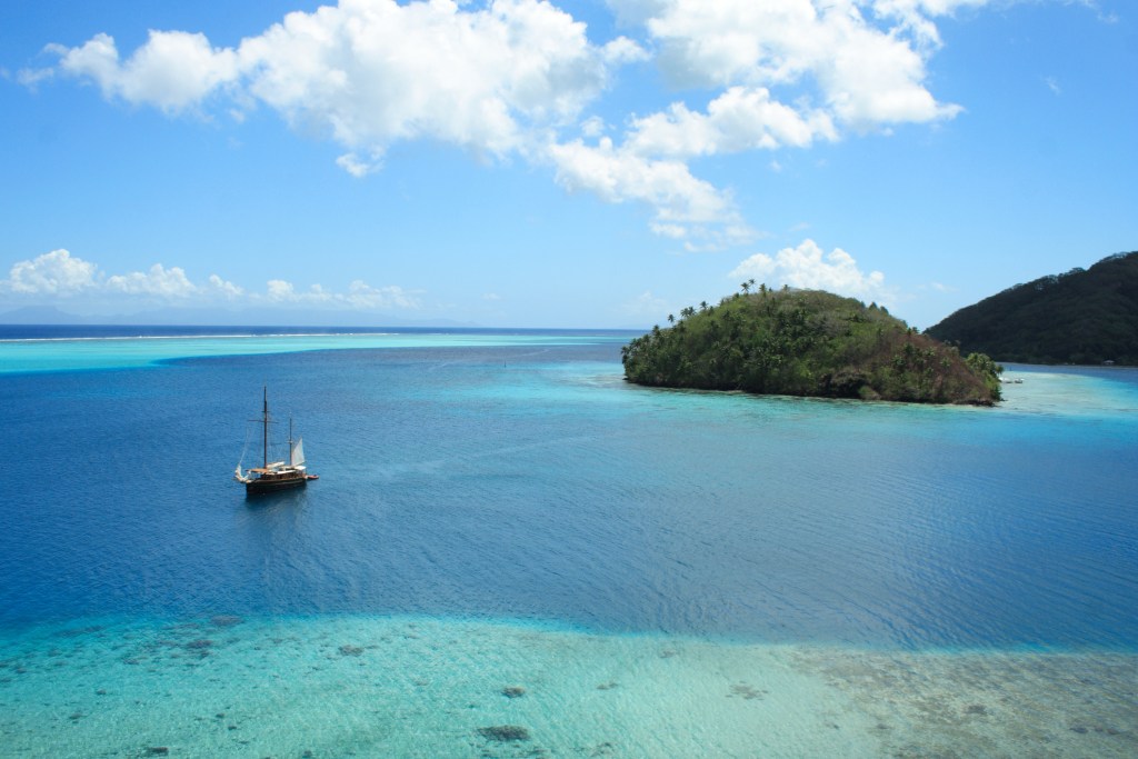 A small tropical island surrounded by bright turquoise water with a lone sailboat.
