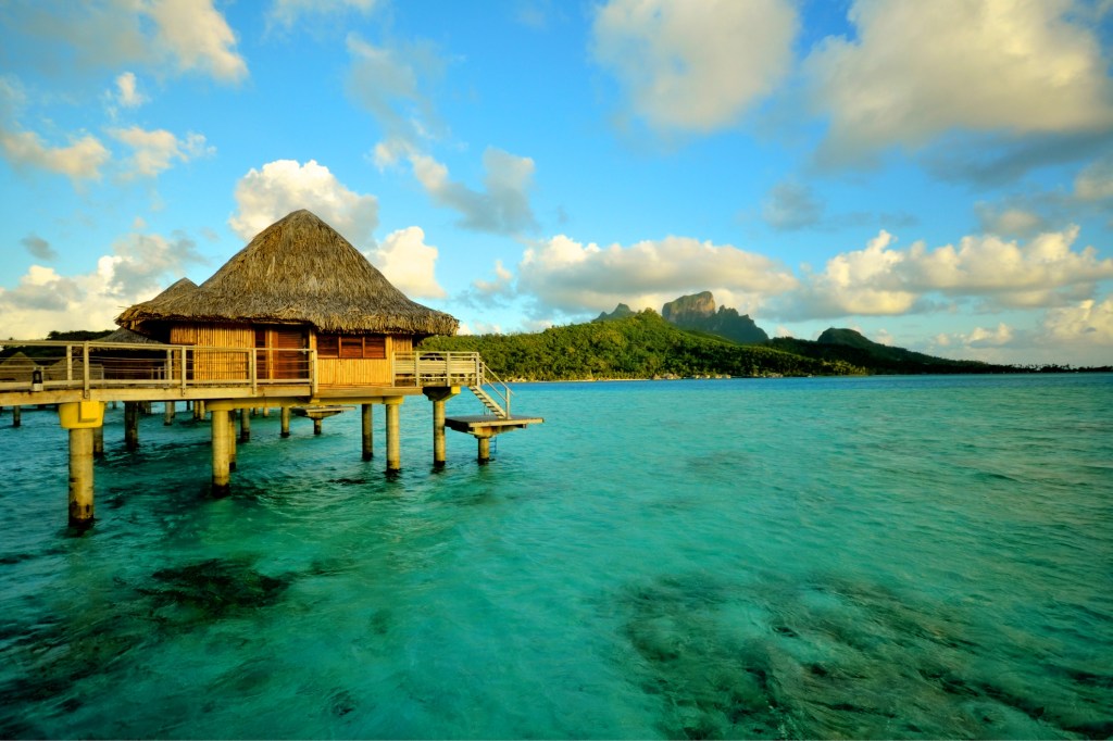 A famous overwater bungalow with a thatched roof sits over turquoise water with a tropical island in the background.