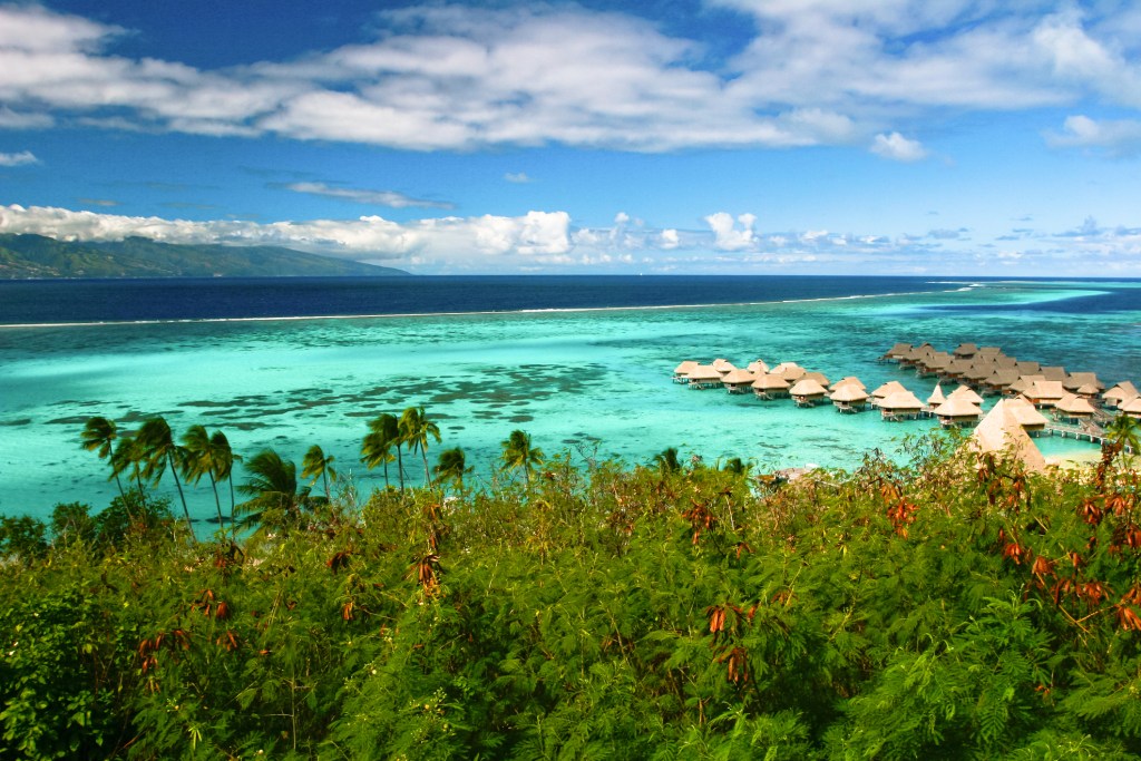 An aerial view of a turquoise lagoon with clusters of overwater bungalows and lush green rainforest in the foreground.