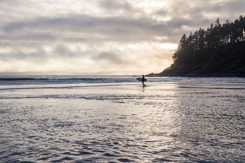 Image of surfer walking on beach with grey clouds reflecting in water.