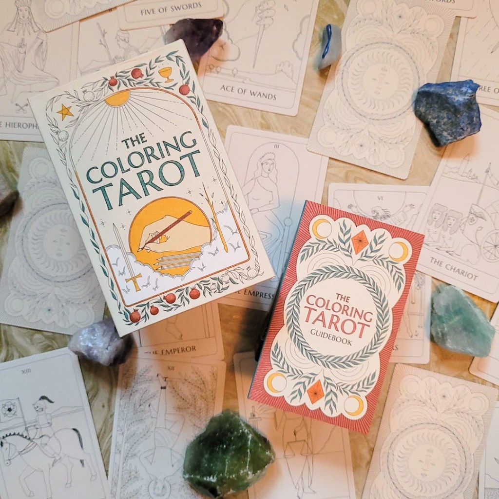 Photo of "The Coloring Tarot" box and guidebook laid above cards from the deck