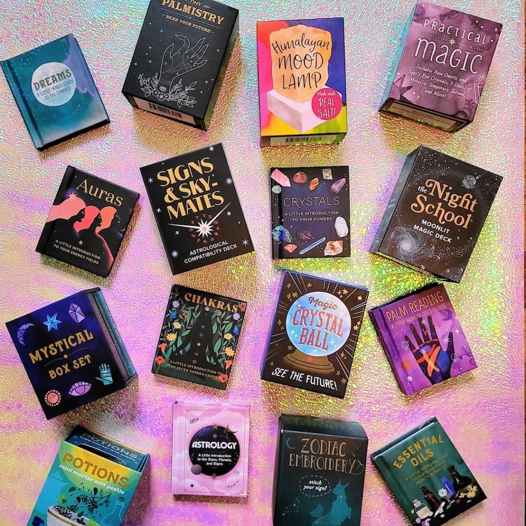 Photo of various RP Mystic mini book, deck, and kit offerings set on a pink and yellow iridescent backdrop