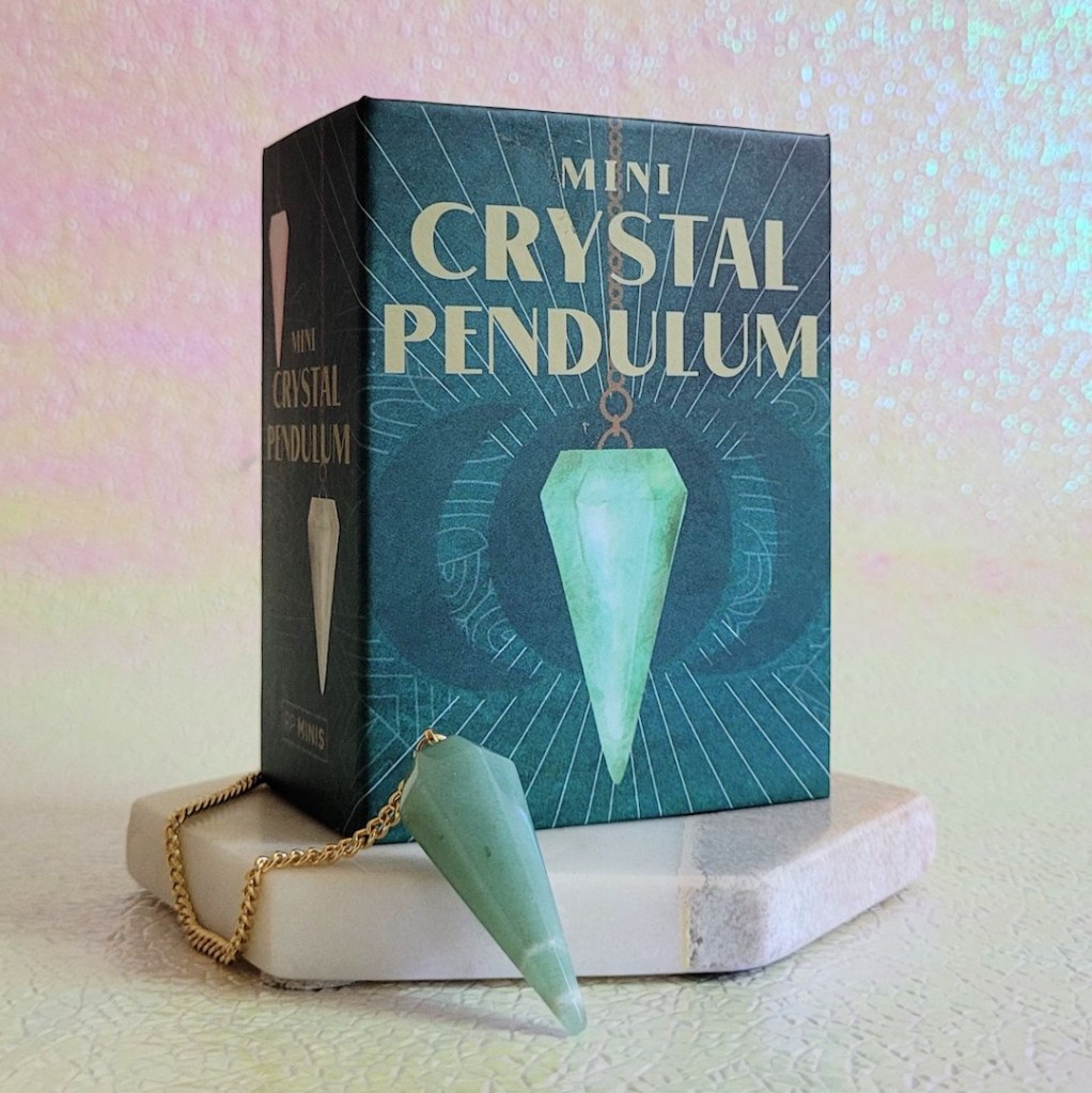 Photo of the "Mini Crystal Pendulum" box and crystal standing against a pink and yellow iridescent background