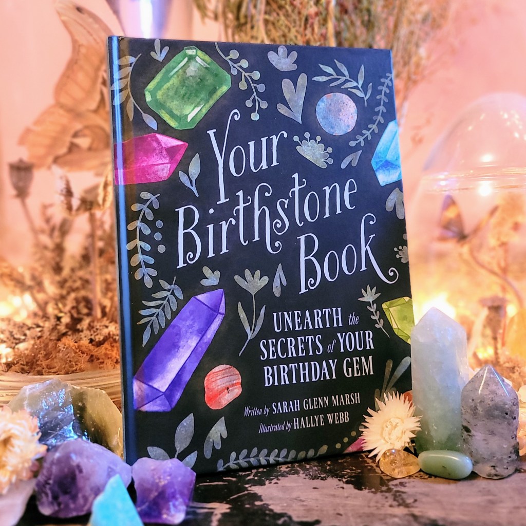 Photo of "Your Birthstone Book" standing among various crystals and floral decor against a softly lit pink background