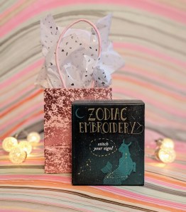 Photo of the "Zodiac Embroidery" box in front of a mini gift bag filled with tissue paper surrounded by tiny lights, against a marbled background