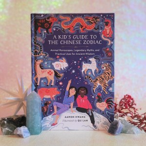 Photo of “A Kid’s Guide to the Chinese Zodiac” standing among crystals, flowers, and a decorative white starburst, in front of a pink and green iridescent background.