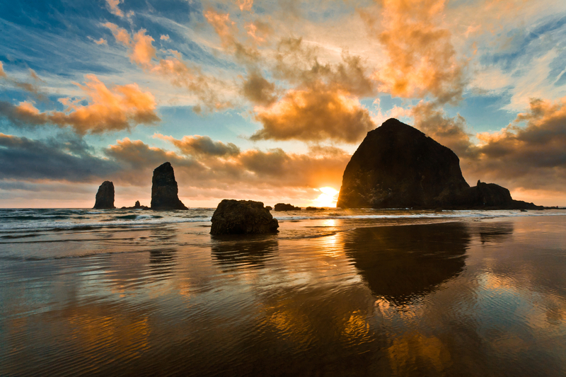 Image of golden cloudy sunset over rock formations reflected on beach.