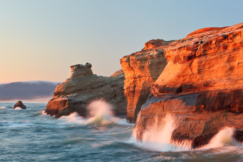 Waves crash onto rocky cliffs glowing under orange sunset.