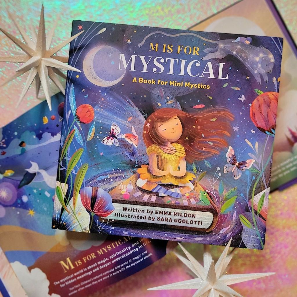 Photo of a closed copy of “M is for Mystical” laid above an open copy of the same book. Both are laid next to decorative white starbursts above a pink and green iridescent background.