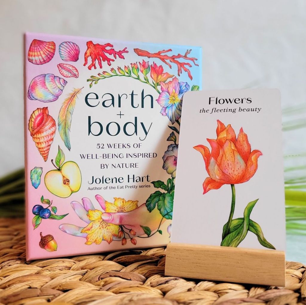 Photo of the “Earth + Body: 52 Weeks of Well-Being Inspired by Nature” keepsake box standing next to a card from the deck sitting in the included wooden stand. The box and stand are standing on a woven mat in front of the stems of a bouquet.