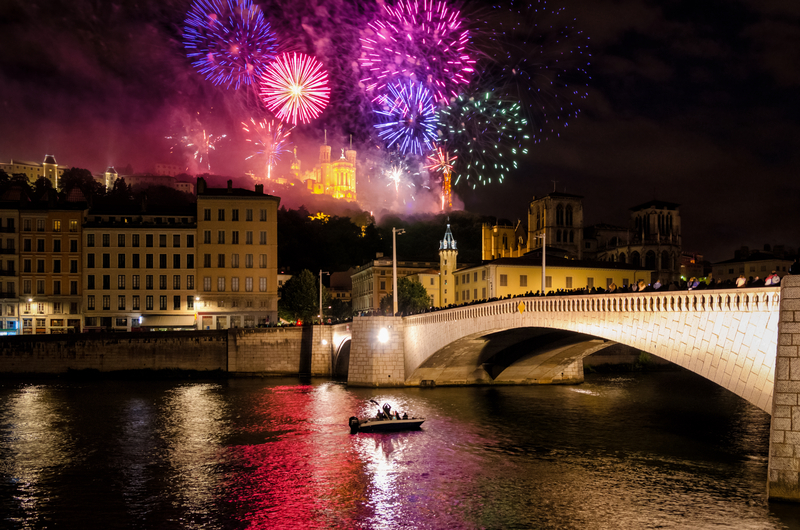 Colorful pink and purple fireworks explode in the background of stone buildings along a reflective river with a beautiful stone bridge.