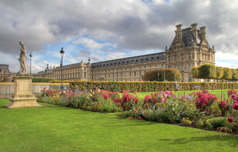 Image of green manicured garden with statues with low stone building of the Louvre in the distance.