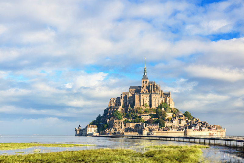 A gothic, stone abbey on an island rising from marshy water and green grass under cloudy blue sky