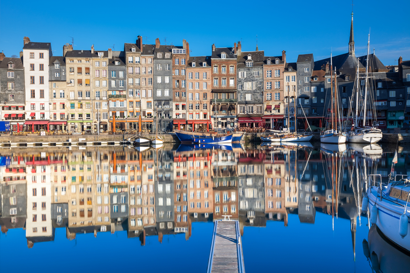 Narrow, colorful rowhouses and boats reflected in calm harbor under bright blue sky.