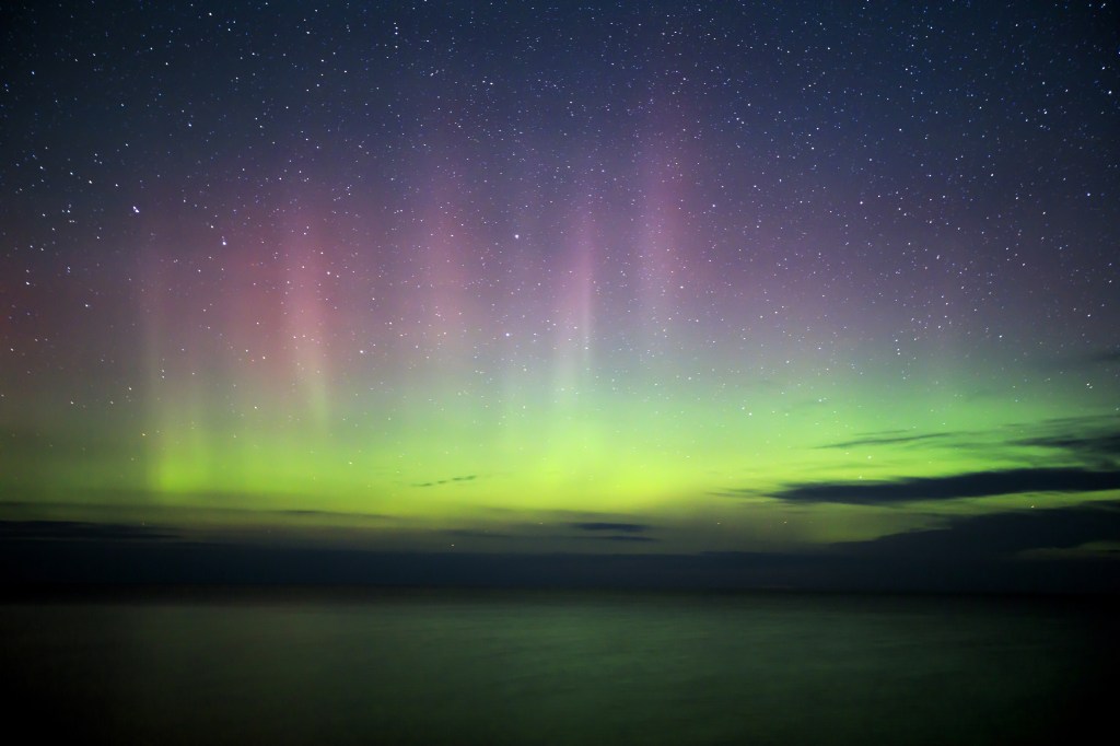 green and pink northern lights hovering low, scattered with dark splotches of clouds over lake superior