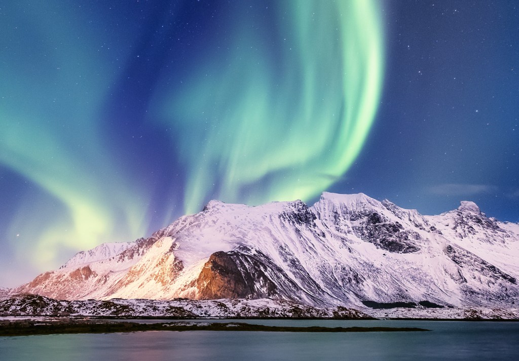 pale green northern lights filling the inky blue night sky above Norway, over a massive snowy mountain and calm lake in the foreground