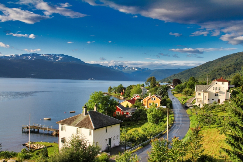 Village buildings perched on lush hillside overlooking calm fjord under deep blue sky