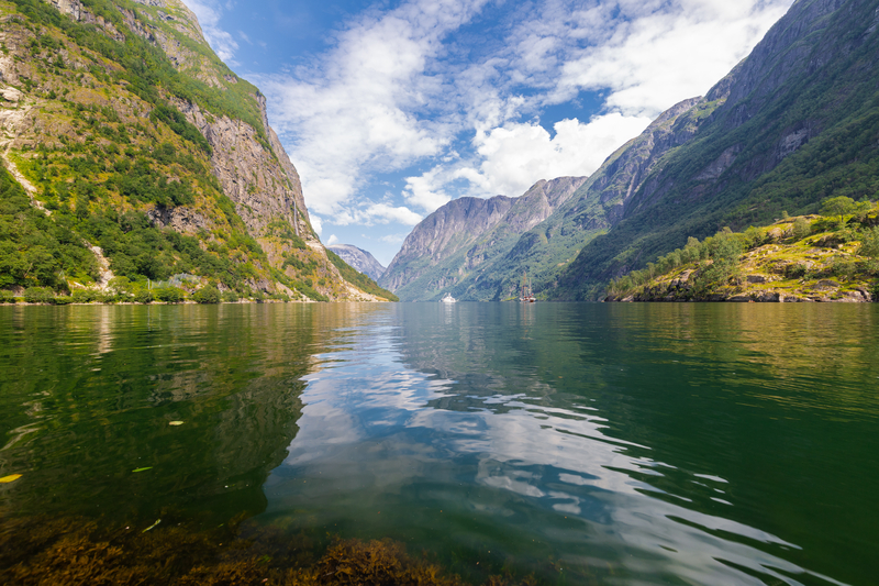 View of the entrance to a fjord with tall mountains rising on either side and clouds reflected in the water