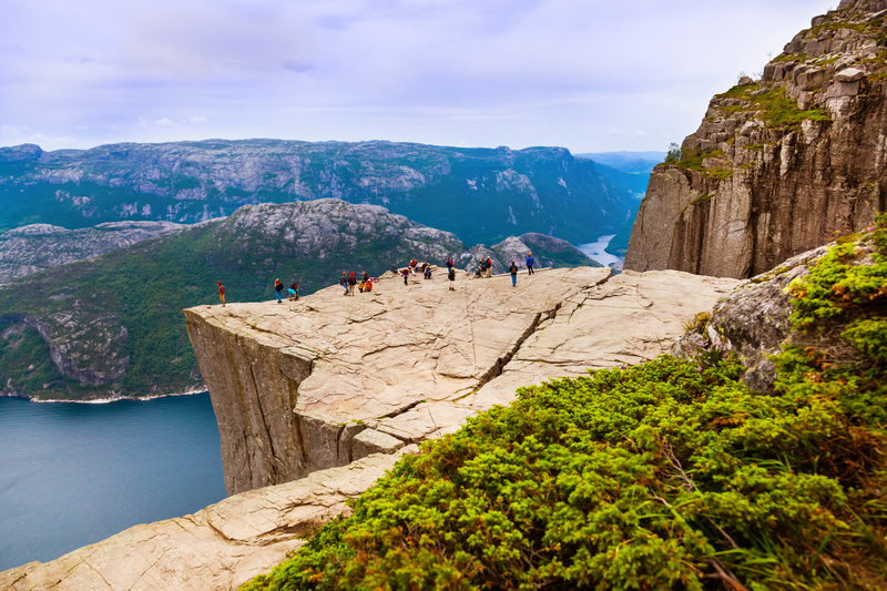 Small group of people standing on top of flat rock cliff overlooking fjord