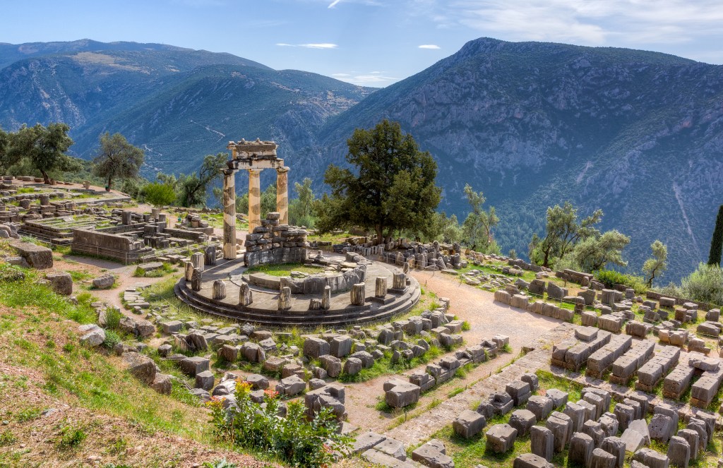 a crumbled ancient temple with three columns still holding up a portion of the roof sits atop a sunny mountain side