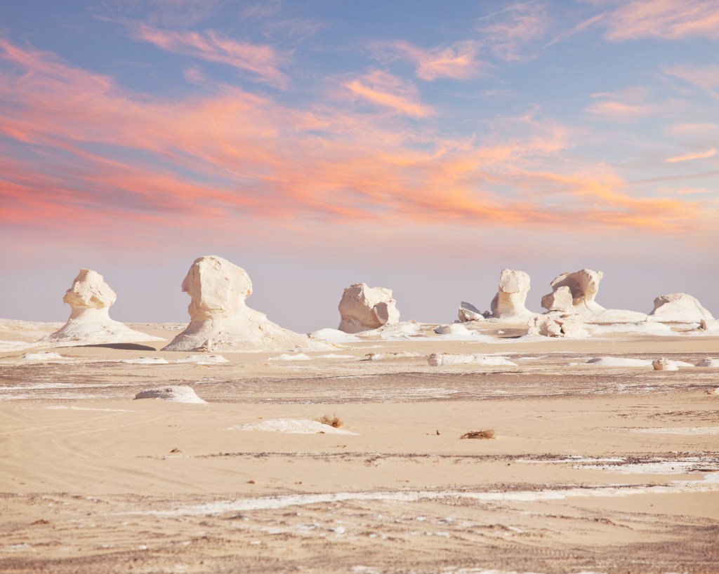 bulbous white stones jut out of the sandy desert floor during sunrise with soft pink and orange clouds in the sky