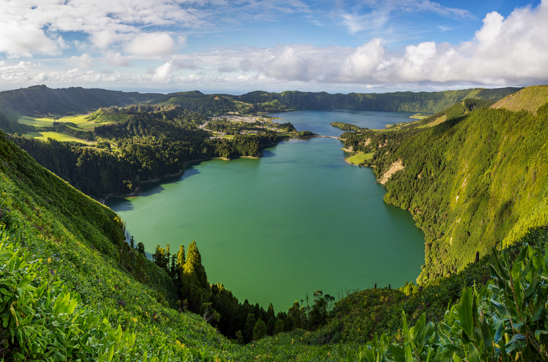 A blue-green lake surrounded by towering lush green mountains as seen from a clifftop viewpoint