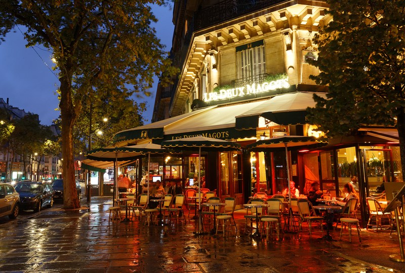 A lit-up cafe with tables and chairs on the patio on a rainy evening