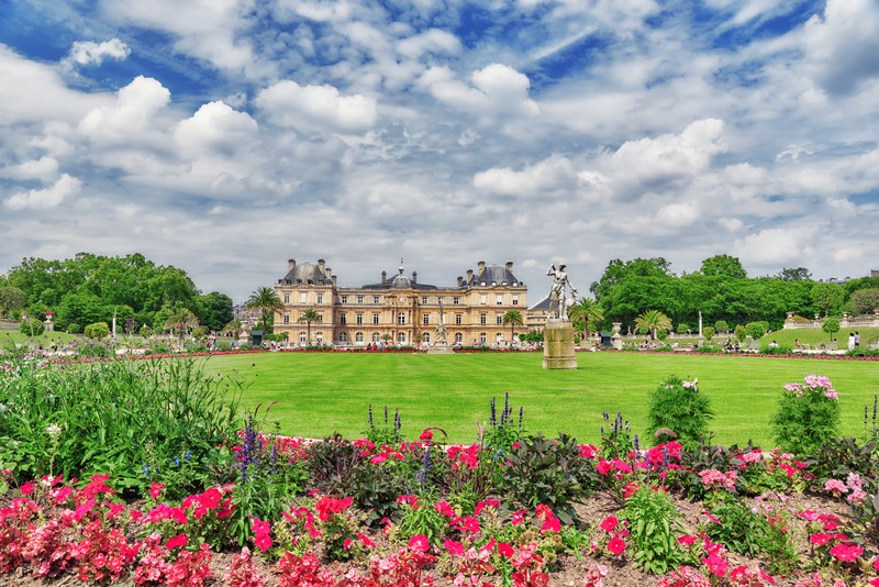 A large green lawn and colorful red and pink flower gardens with stately building in the distance under cloudy blue sky.