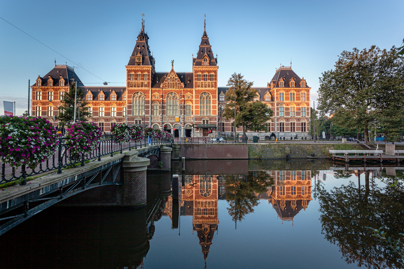 An ornate brown brick building with white trim reflected in a serene canal at sunset